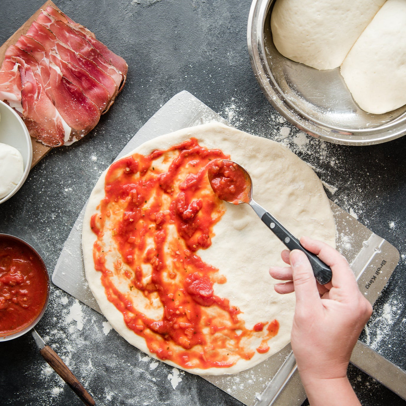 Person spreading tomato sauce on pizza dough with ingredients around on a dark surface