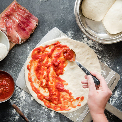 Person spreading tomato sauce on pizza dough with ingredients around on a dark surface